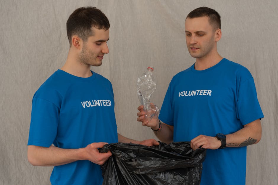 Two male volunteers wearing blue t-shirts with the word 'VOLUNTEER' printed on the front are participating in a recycling or community clean-up activity indoors. One volunteer is holding a black garba