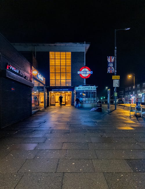 This image shows the exterior of Harringay Green Lanes station at night, illuminated by warm lighting from the station entrance and surrounding streetlights. The urban scene features a paved sidewalk 