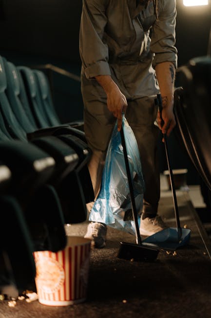 A sanitation worker dressed in bright orange high-visibility clothing, including a hooded jacket and pants, is sweeping a paved sidewalk with a red-bristled broom. The worker is using a blue dustpan, 
